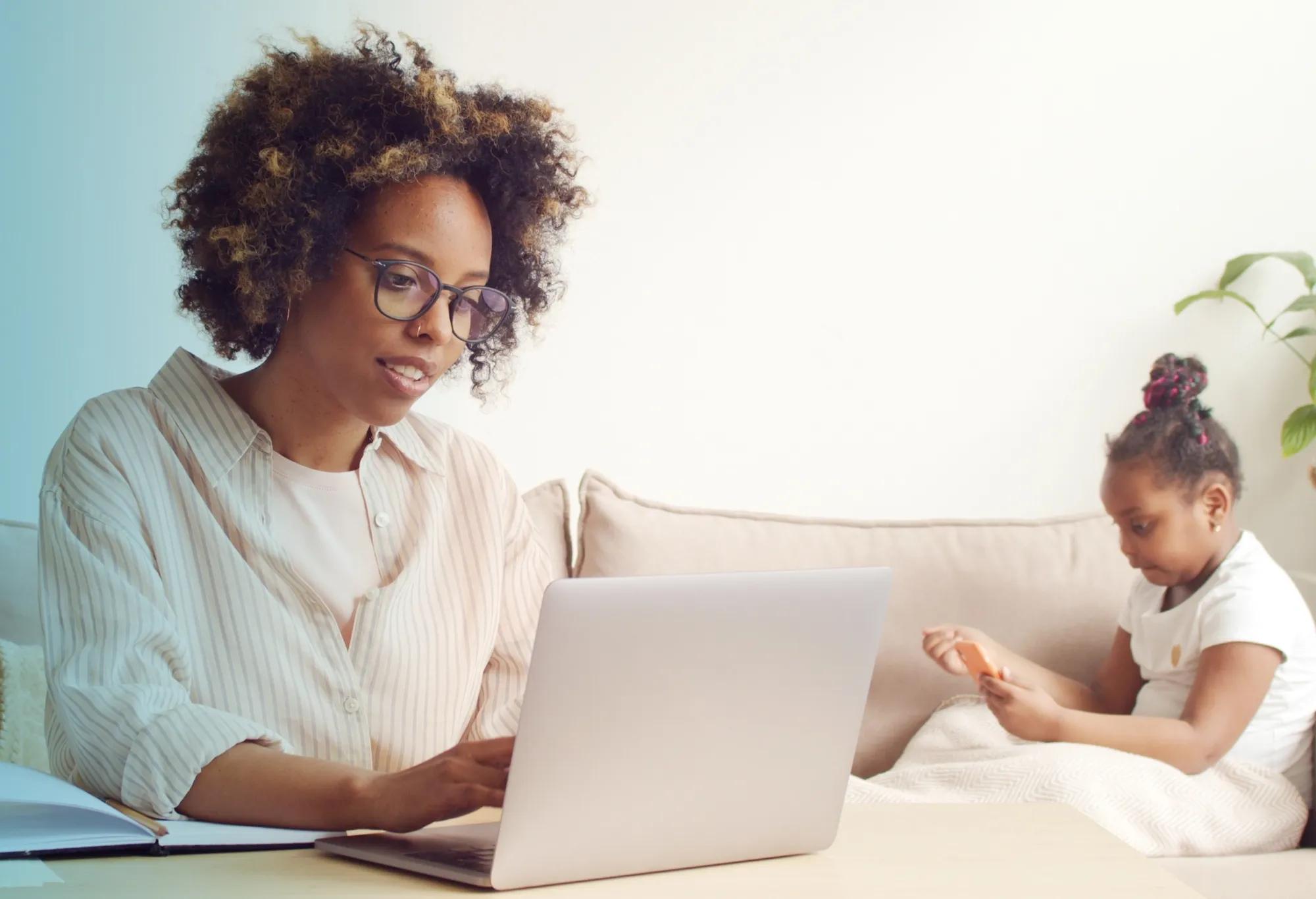 An adult learner taking notes from her online healthcare training program while her child is on the couch in the background