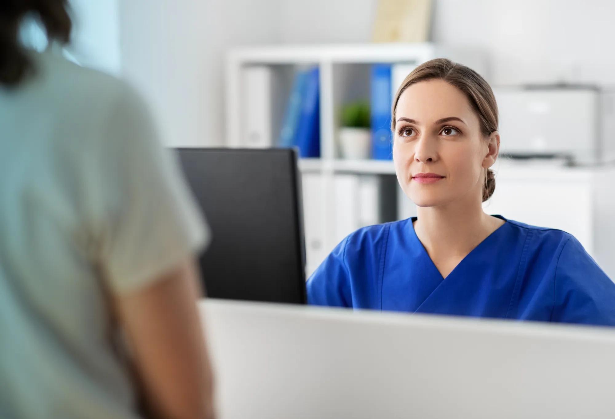 Woman in an allied healthcare role, at the front desk of a healthcare institution, helps a patient.