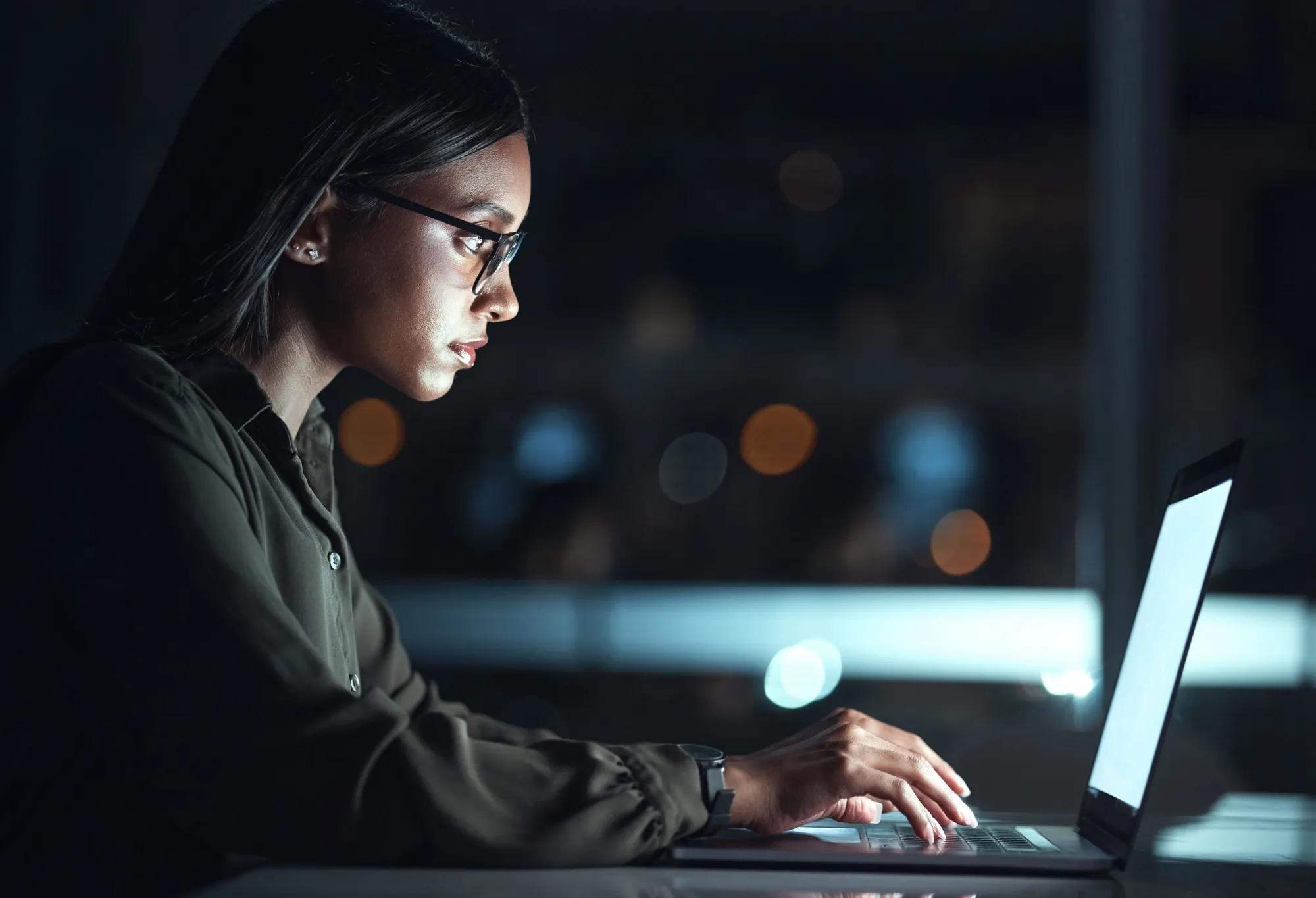 An adult student doing schoolwork on a laptop at night with no room lights on, so their face and front upper body are illuminated by the light from the laptop screen.