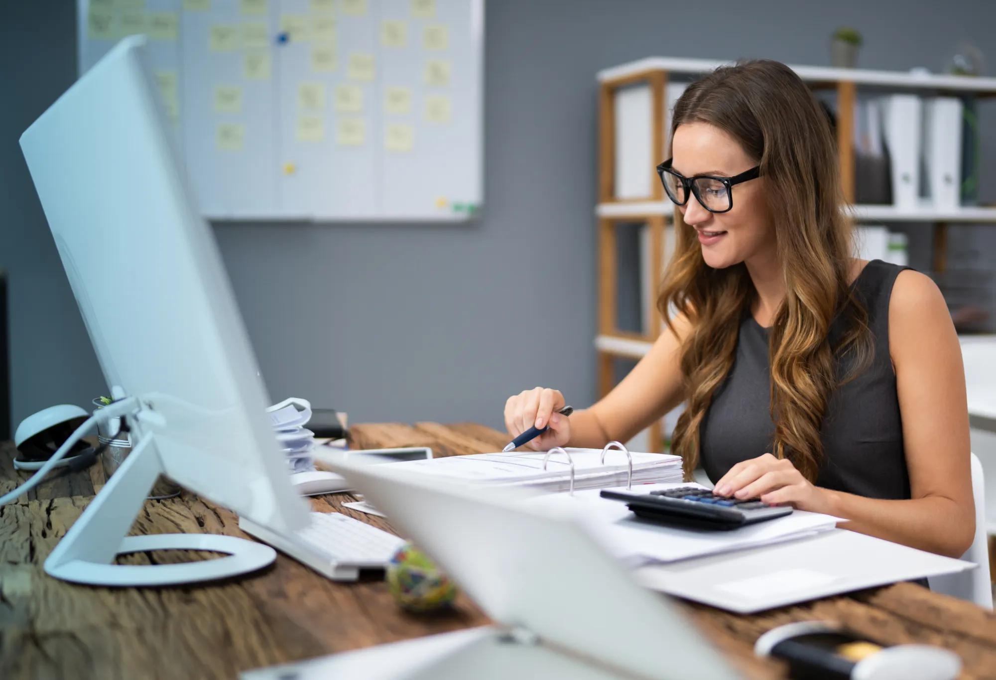 A medical biller and coder working at a desk with a calculator, notebook, and computer