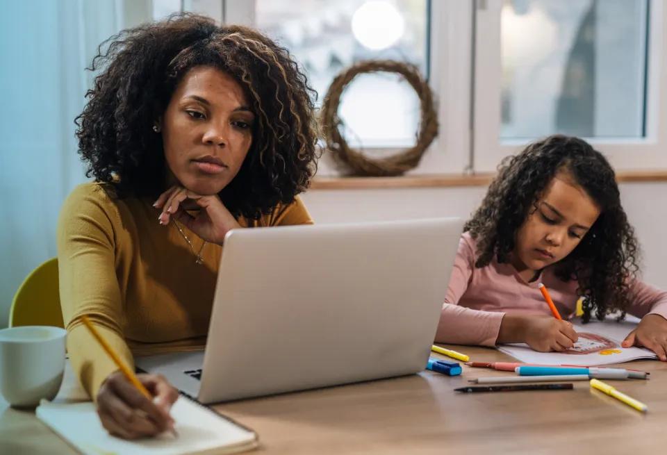 an adult learner working on a laptop with a child nearby as she tries to balance home, work, and school while earning a healthcare degree
