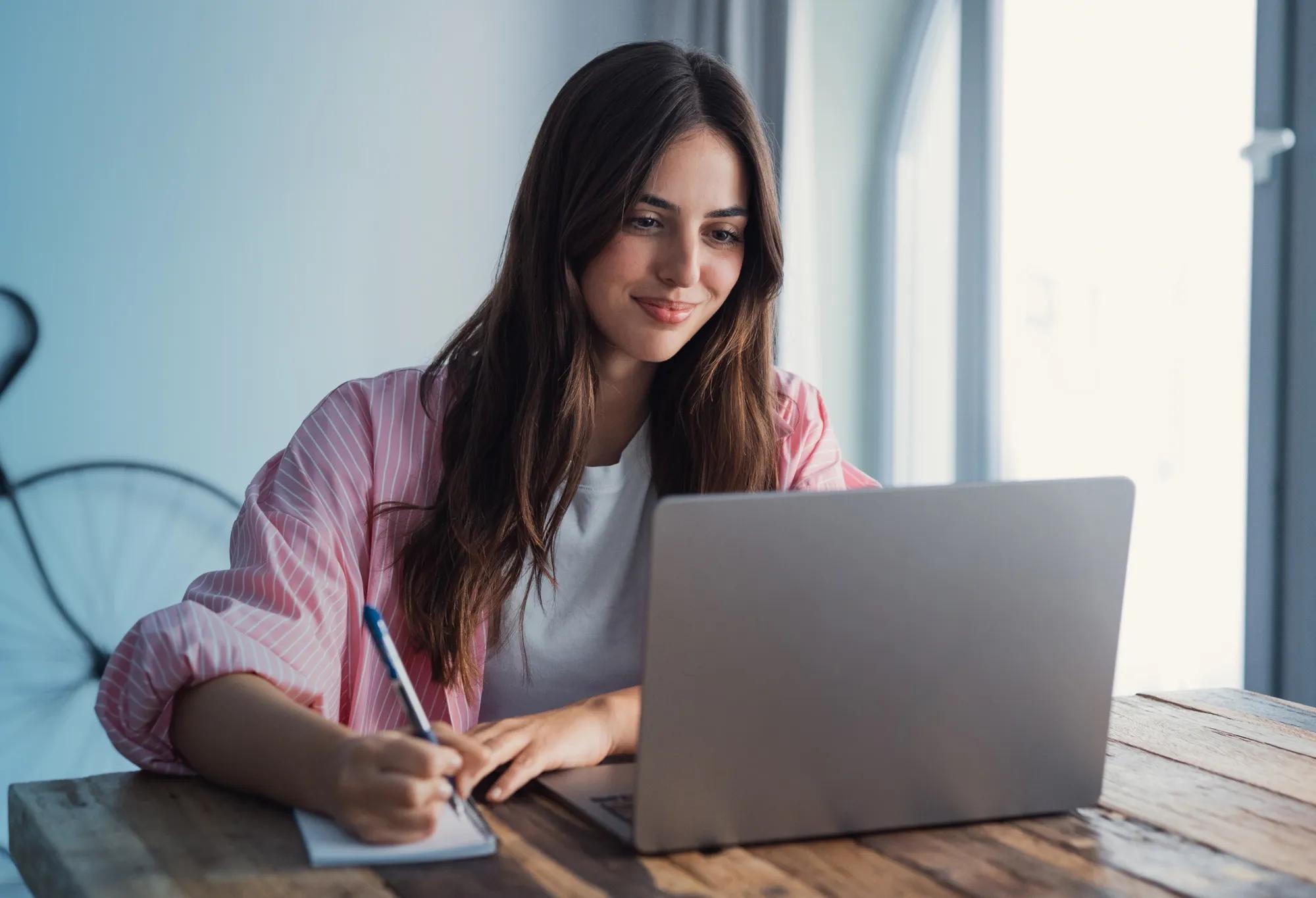 A woman sitting at a table with a laptop and writing notes on a tablet while doing online schoolwork
